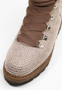 Bota con cordones de ante en rosa claro, adornada con tachuelas plateadas. Presenta cordones marrones y una suela de goma resistente con un borde cosido.