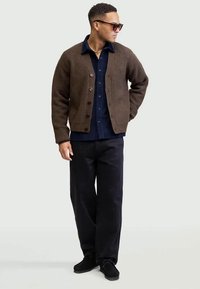 Uomo che indossa occhiali da sole, cardigan marrone con bottoni, camicia blu navy, pantaloni neri e scarpe nere, in piedi con una mano in tasca.
