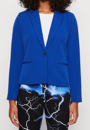 Femme portant un blazer bleu ajusté sur un top blanc et un pantalon noir avec un motif éclair bleu et blanc, debout devant un fond uni.