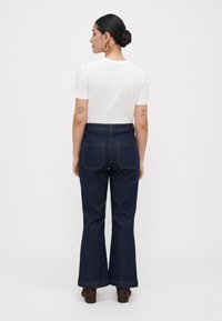Kvinna med uppsatt hår i en knut, som bär örhängen i form av ringar, en vit kortärmad skjorta, mörka utsvängda jeans och bruna skor, står vänd bort.