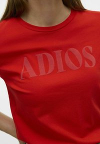 T-shirt en coton rouge avec un grand imprimé semi-transparent "ADIOS" dans une couleur contrastante, col rond et manches courtes.