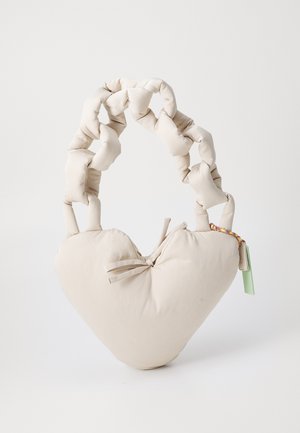 Bolsa em forma de coração, almofadada, em tecido beje claro, com uma alça grossa trançada e uma etiqueta colorida como detalhe. Detalhe de laço amarrado na parte da frente.