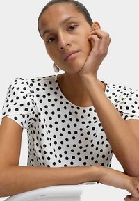Weiße Bluse mit schwarzen Polka-Dots, kurzen Puffärmeln, glatter Textur und rundem Ausschnitt. Das Model stützt ihr Kinn auf eine Hand.