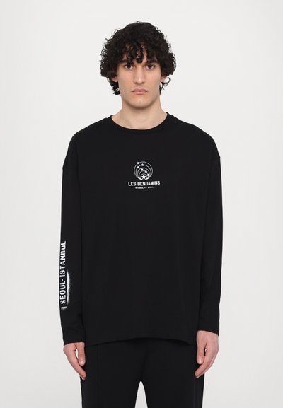 T-shirt noir à manches longues en coton, avec un logo blanc sur la poitrine et le texte "SEOUL-ISTANBUL" le long de la manche gauche.