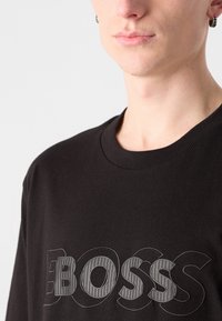 Juodos medvilnės marškinėliai su dideliu, tekstūriniu pilku "BOSS" logotipu priekyje. Dizainas apima klasikį apval�ų kaklą ir trumpas rankoves.