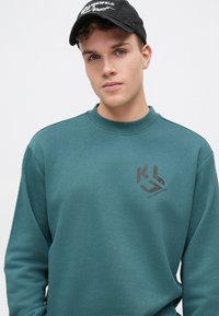 Turkis genser i myk stoff med rund hals og lange ermer. Har en brun logo på brystet. Caps med logo.