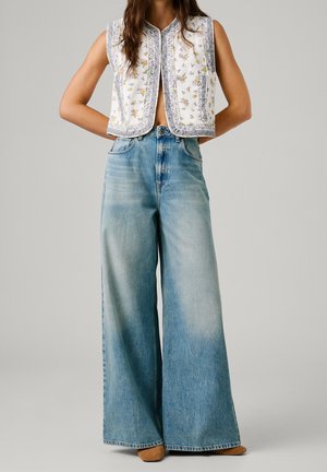 Kobieta ubrana w kwiecistą bezrękawnik i wyblakłe, niebieskie dżinsy z wysokim stanem i szerokimi nogawkami, z brązowymi butami, stojąca na jednolitym tle.