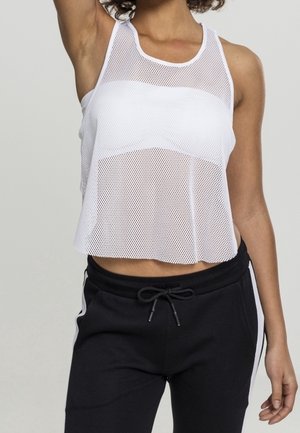 Femme portant un débardeur en maille blanche par-dessus un soutien-gorge de sport blanc et un pantalon noir à cordon avec des bandes blanches sur les côtés.