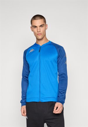 Giovane uomo che indossa una giacca sportiva blu con zip, maniche blu scuro e pantaloni neri, posando davanti a uno sfondo grigio uniforme.