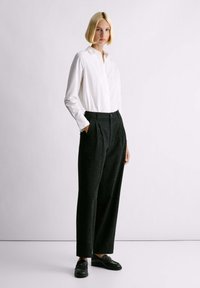 Camicia bianca a maniche lunghe con dettagli a bottoni, abbinata a pantaloni neri plissettati con texture e mocassini neri. Design minimalista e vestibilità rilassata.