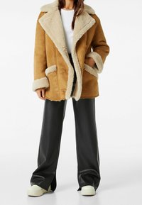Hnědý kabát z ovčího rouna s béžovým kožíškem na límci a manžetách, dvěma předními kapsami a volným střihem, zkombinovaný s černými kalhotami připomínajícími kůži a teniskami.