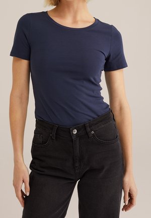 Kobieta ubrana w dopasowany granatowy T-shirt z krótkim rękawem, wsunięty w czarne, wysokie jeansy na tle jednolitego beżowego tła.