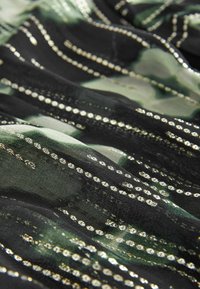 Tissu noir avec des motifs floraux verts et des rangées de petites perles dorées brillantes formant des motifs linéaires à la surface.