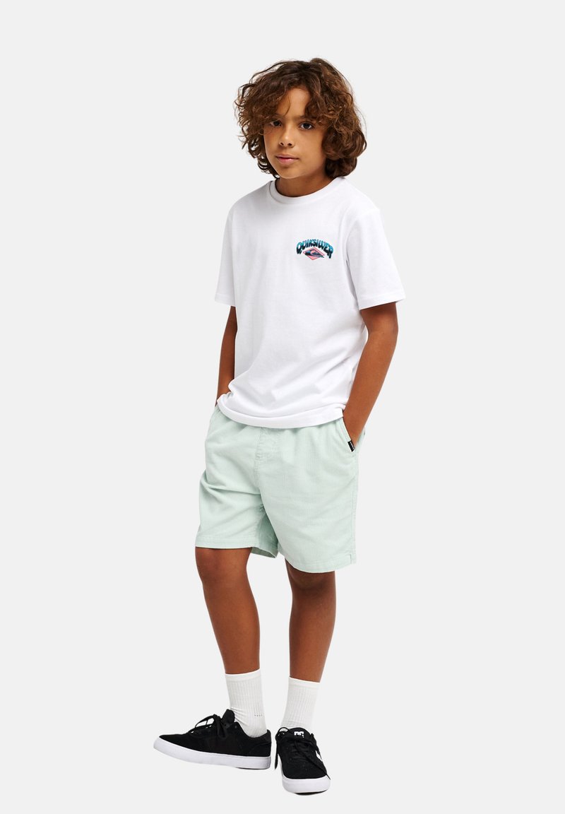 Αγόρι με σγουρά μαλλιά, φορώντας λευκό μπλουζάκι με σχέδιο, ανοιχτό πράσινο σορτς, λευκές κάλτσες και μαύρα αθλητικά παπούτσια, στέκεται με τα χέρια στις τσέπες.