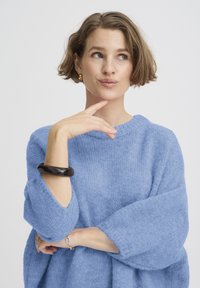 Pull en maille bleu clair avec manches larges. Le modèle porte un bracelet noir et des boucles d'oreilles en or, avec une finition subtilement texturée.