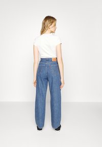 Calças de ganga azul de perna larga com uma cintura alta, apresentando dois bolsos traseiros e um rótulo da marca visível. Combinadas com uma blusa branca de manga curta.