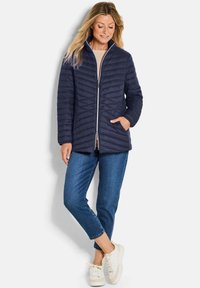 Marineblauwe gewatteerde jas met hoge kraag, frontale rits, twee zakken aan de zijkant, gecombineerd met blauwe jeans en witte sneakers. Gladde textuur.