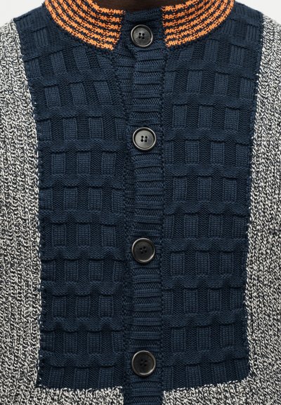 Πλεκτή ζακέτα με μπροστινή όψη σε ναυτικό μπλε με τέσσερα μαύρα κουμπιά, μανίκια σε γκρι και λευκή απόχρωση με μάρλ υφή, και γιακά με πορτοκαλί ρίγες.