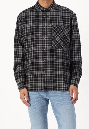 Homme portant une chemise à carreaux noire et grise avec une poche poitrine, associée à un jean bleu clair, debout devant un fond blanc.