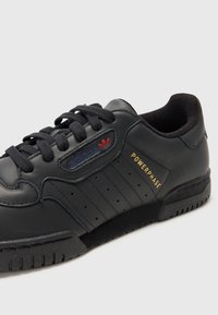 Svart lærsneaker med rund tå, tre perforerte striper, gulltekst og rødt logo-detalj på tungen. Teksturert såle for grep.