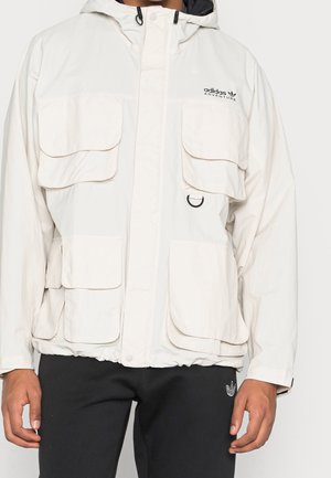 Osoba ubrana w białą kurtkę Adidas Adventure z wieloma dużymi kieszeniami i czarnym kapturem na sznurkach, w połączeniu z czarnymi spodniami Adidas.