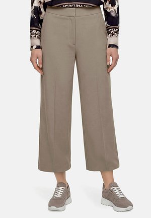 Femme portant un pantalon large cropped beige, un haut à imprimé animalier et des baskets grises, debout devant un fond uni.