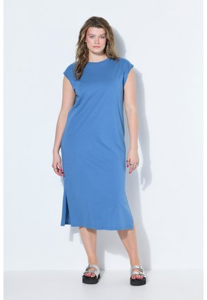 Femme debout portant une robe midi bleue sans manches avec une fente latérale et des sandales à boucle argentée sur fond blanc.