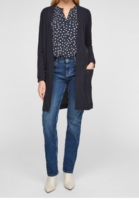 Marinblå cardigan med öppen front, långa ärmar och ficka. Vit blus med prickar under, tillsammans med blå raka jeans och beige ankelstövlar.