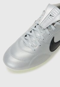Silver atletikskor med en slät läderöverdel, texturerade sidor, platta snören och en svart swoosh-logotyp. Har en perforerad detalj.