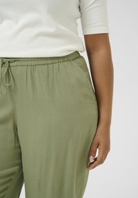 Light olive green, loose-fitting pants with an elastic waistband and drawstring. Soft, smooth fabric with side pockets.