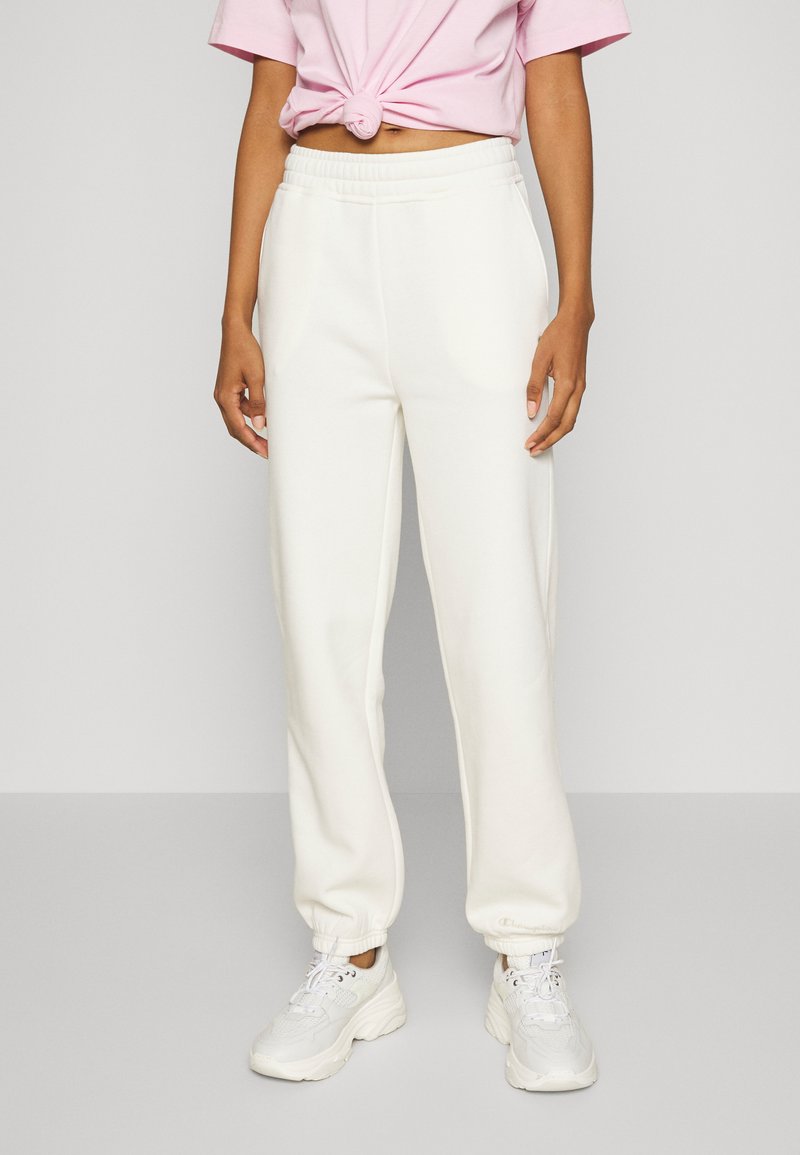 Kvinne iført hvite joggebukser med elastisk midje, hvite sneakers og en knyttet rosa crop topp mot en enkel grå bakgrunn.