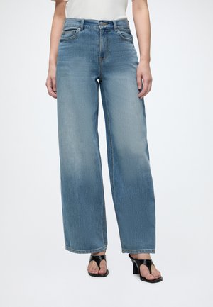 Vrouw draagt blauw, hoog geplaatste, wijd uitlopende jeans gecombineerd met zwarte open-toe sandalen met hak tegen een effen achtergrond.