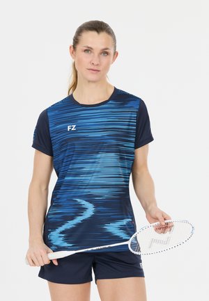 Kvinde iført blåt mønstret sportstøj, der holder en hvid badmintonketcher mod en ensfarvet baggrund.