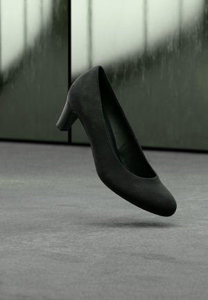 Zapato de tacón medio de ante negro suspendido sobre suelo alfombrado gris con paneles de vidrio esmerilado al fondo.