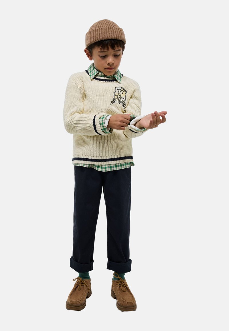 Pull en maille crème avec des rayures bleu marine et noires, superposé à une chemise à carreaux verts. Pantalons foncés et chaussures marron avec des semelles texturées.