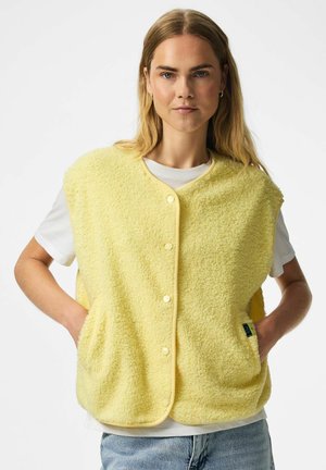 Žena s dlhými blond vlasmi nosí žltú fleecovú vestu na gombíky cez bielu košeľu a modré džínsy, ruky v vreckách vesty, neutrálne pozadie.
