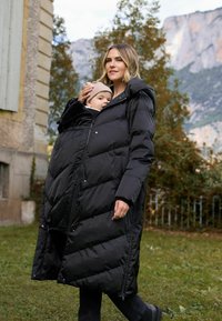 Manteau noir gonflé conçu pour porter un enfant, doté d'une fermeture éclair à l'avant, d'une grande capuche et de poches latérales. Texture rembourrée avec motif en chevrons.