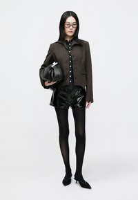 Bruine jasje met krijtstreep, zwart overhemd met knopen, glanzende zwarte korte broek, zwarte panty's en puntige zwarte hakken. Houdt een zwart gestructureerde tas vast.