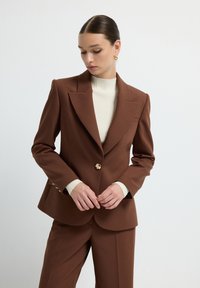 Blazer marrone su misura con rever, dettagli in bottone dorato e spalle strutturate, abbinato a un dolcevita rigato color crema.