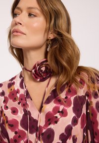 Kobieta o falowanych jasnobrązowych włosach nosi złote kolczyki i kwiecistą bluzkę w odcieniach różu, fioletu i pomarańczu oraz pasujący do niej choker z tkaniny w formie róży.