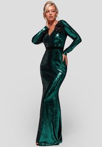 Grøn palietkjole med lange ærmer, V-udskæring, kropsnær silhuet og et sort blomsterornament på den ene skulder. Glat tekstur og hel længde design.