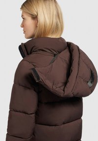 Brun oppustelig jakke med høj krave, stor hætte og synlige syninger. Har lynlås og velcro-detaljer til lukning og justering.