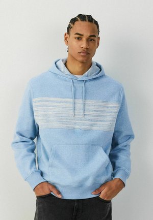 Young man with braided hair wearing a light blue hoodie with horizontal white and blue stripes, hands in pockets, neutral background.