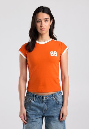 Jonge vrouw met lang donker haar, gekleed in een oranje cropped T-shirt met witte bies en het nummer 89, gecombineerd met blauwe spijkerbroek.