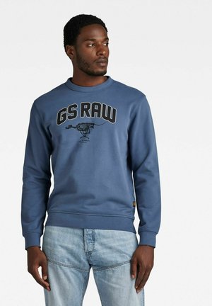 Sudadera azul con cuello redondo, que presenta el texto "GS RAW" y una impresión gráfica. Tejido suave, mangas largas y puños de canalé. Se combina con jeans claros.