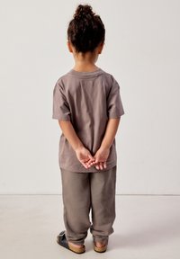 Bambino in piedi, rivolto verso un muro bianco, con le mani incrociate dietro la schiena, che indossa una maglietta marrone, pantaloni larghi e sandali neri.