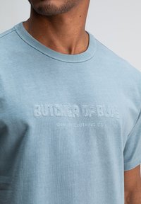 Deel van een persoon die een lichtblauw T-shirt draagt met geborduurde tekst "BUTCHER OF BLUE" en kleinere tekst "QUALITY CLOTHING CO."