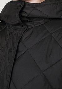 Chaqueta acolchada negra con cuello alto, con patrones de diamante, botones a presión y una textura de tela lisa y brillante.