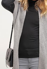 Šedý pletený kardigan s otevřeným předním designem, nošený přes černý rolák. Obsahuje dlouhé rukávy a černou crossbody tašku.