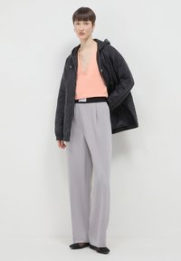 Femme aux cheveux courts portant une veste matelassée noire sur un top corail à col en V, un pantalon large gris clair et des ballerines noires, se tenant contre un mur blanc.